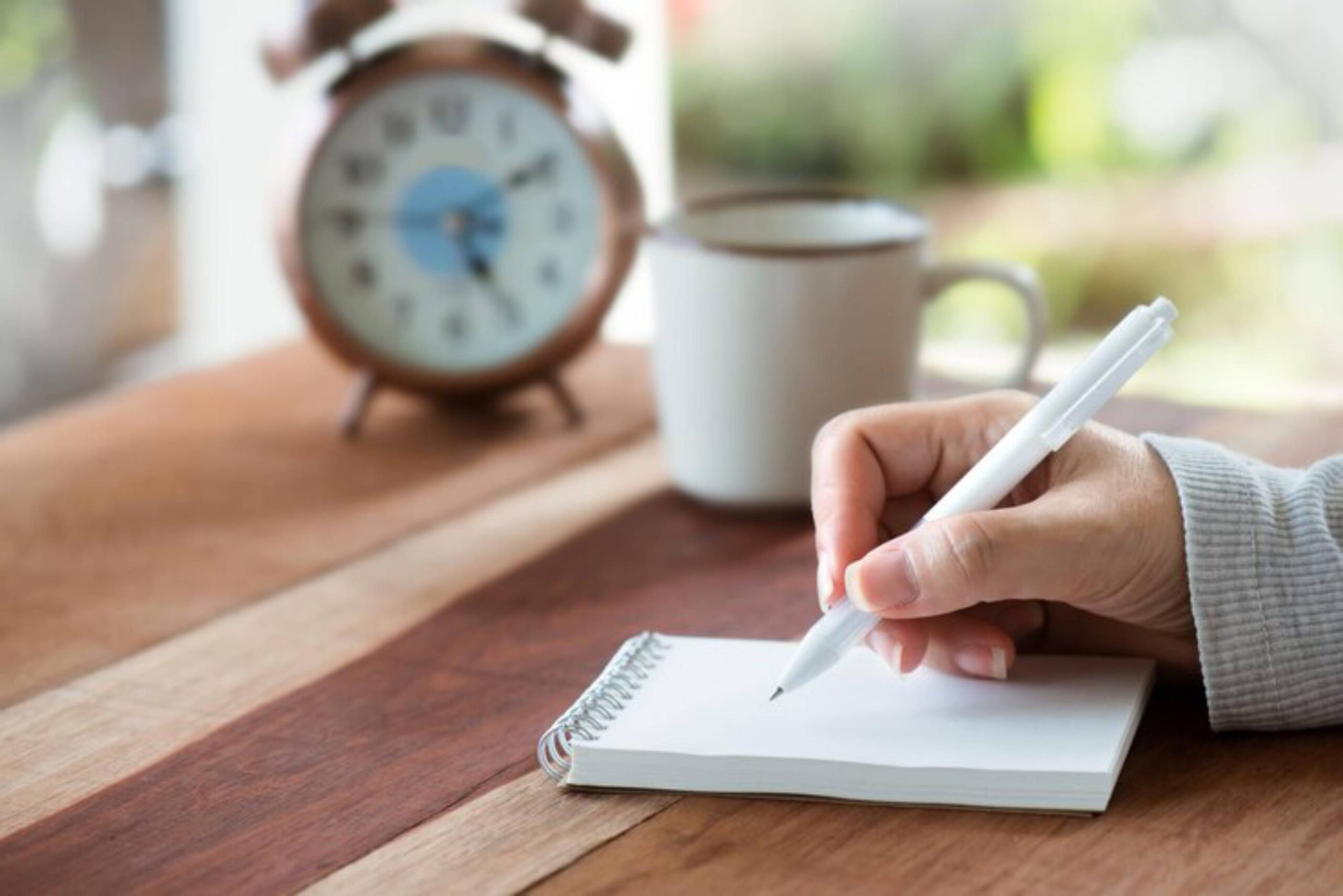 A person writes in a spiral notepad with a white pen, beside a coffee mug and a vintage alarm clock on a wooden table.