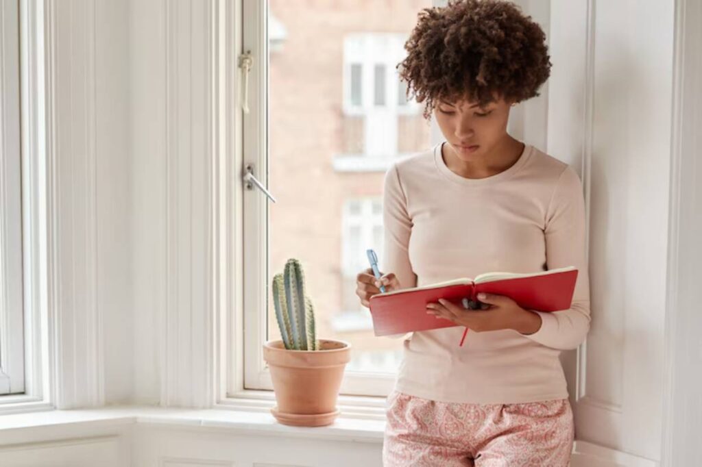 A person stands by a window, writing in a red notebook, with a small cactus in a pot nearby and soft natural light filling the room.