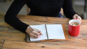 A person writes in an open notebook with a silver pen, holding a red coffee cup on a wooden table.