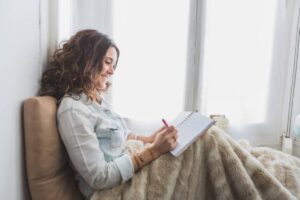 A woman sits comfortably wrapped in a fuzzy blanket, writing in a notebook by a bright window.