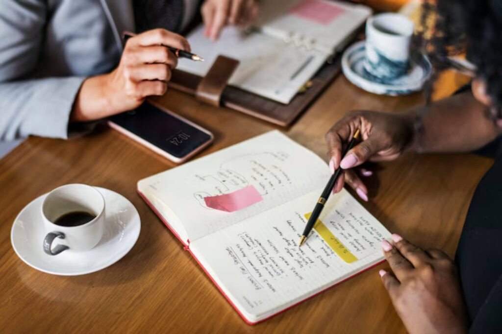 Two individuals are engaged in a meeting at a wooden table, discussing notes in a notebook, with coffee and a smartphone nearby.
