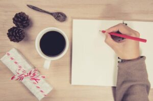 A hand holds a red pencil above a blank notebook, with a cup of coffee, gift, and pinecones nearby on a wooden table.