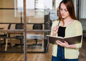 A woman stands with a notebook and pen, deep in thought, in a well-lit office space with rustic decor.