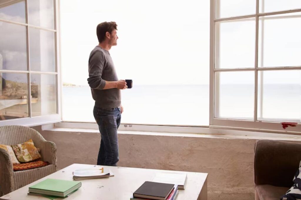 A man stands by a large window overlooking a serene body of water, holding a cup, with cozy furniture surrounding him.