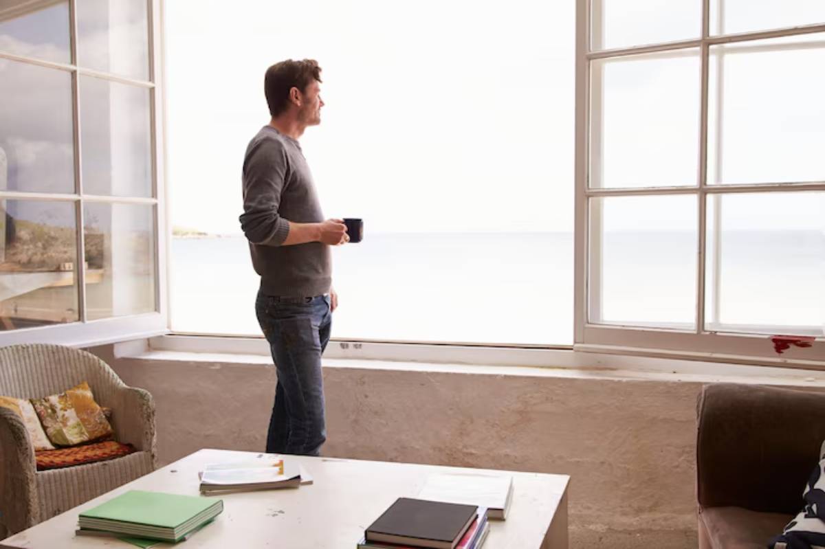 A man stands by a large window overlooking a serene body of water, holding a cup, with cozy furniture surrounding him.