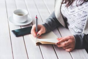 A person writes in a blank notebook with a pen, a coffee cup, and a smartphone on a wooden table.