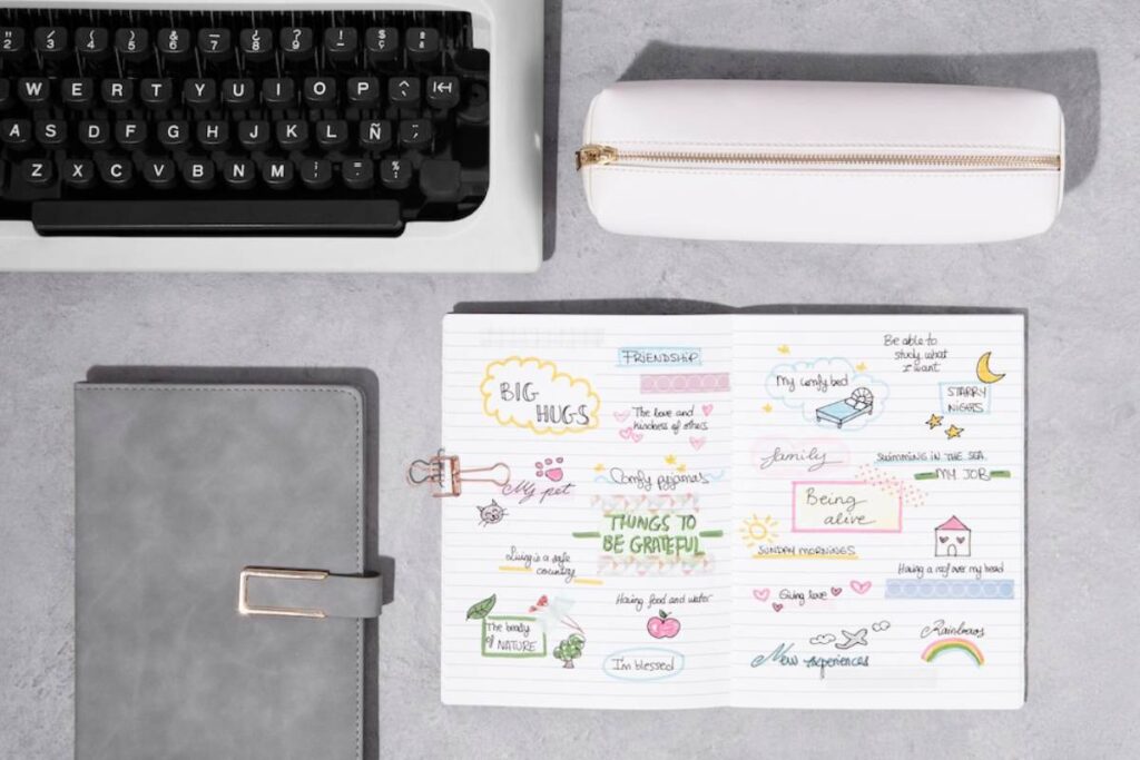 A top-down view of stationery: a typewriter, a gray notebook, and an open journal filled with colorful doodles about gratitude and friendship.