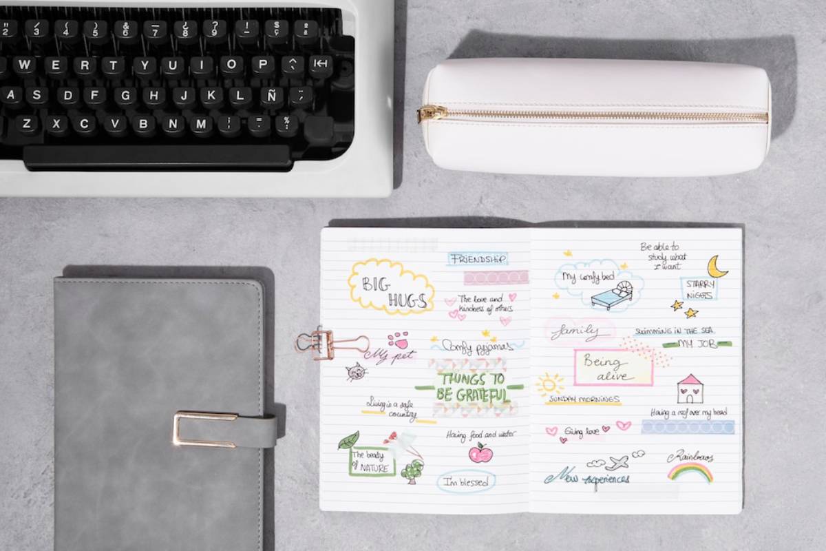 A top-down view of stationery: a typewriter, a gray notebook, and an open journal filled with colorful doodles about gratitude and friendship.