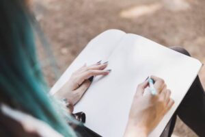 A person with teal hair sits outdoors, holding a pen over an open, blank notebook, poised to write.