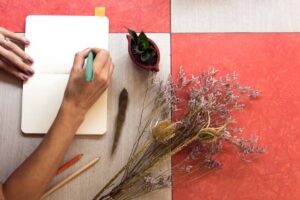 A hand holding a green pen writes in a blank notebook, surrounded by dried flowers, a potted plant, and colored pencils on a textured table.