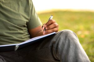 A person sitting outdoors writes in a notebook, holding a pen in their right hand, with grass and blurred background.
