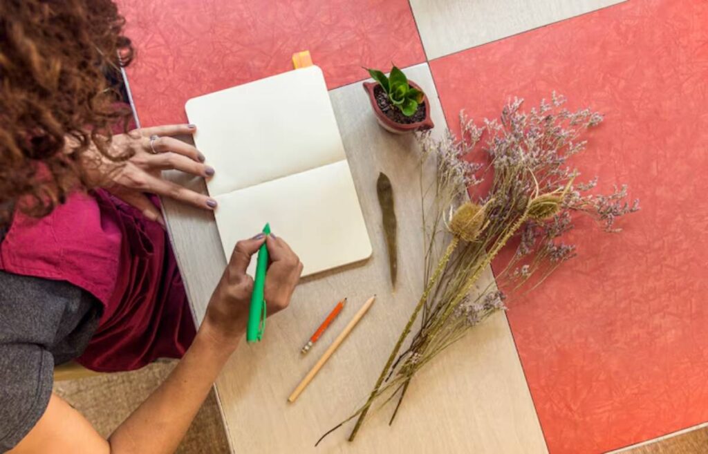 A person with curly hair writes in a notebook with a green pen, surrounded by dried flowers, a small plant, and colored pencils.