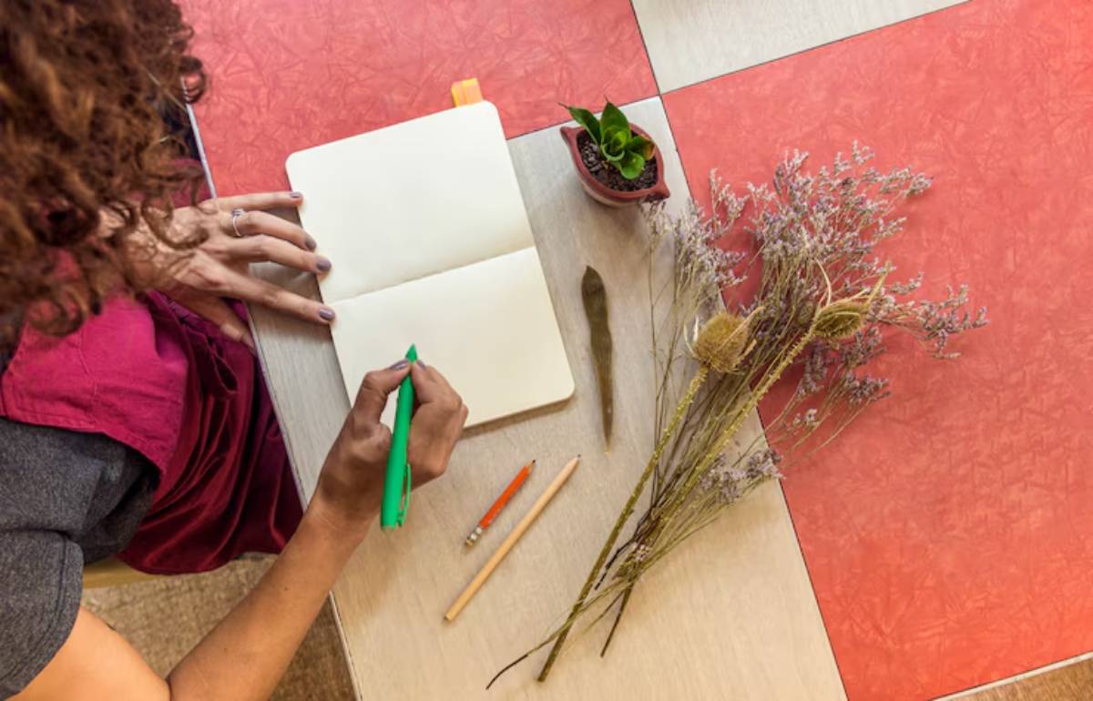 A person with curly hair writes in a notebook with a green pen, surrounded by dried flowers, a small plant, and colored pencils.