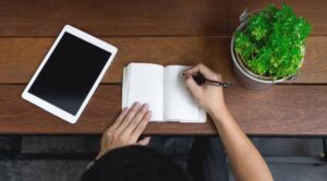 A person writes in a blank notebook while using a tablet, with a small potted plant nearby, all arranged on a wooden table.