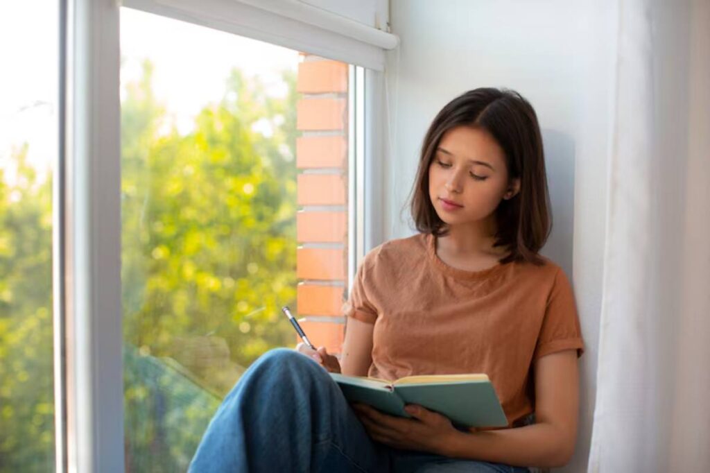 A person sitting by a window, writing in a notebook, surrounded by lush greenery in soft sunlight.