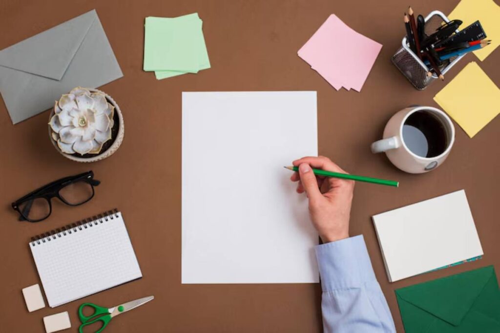 A hand holding a green pencil above a blank paper, surrounded by stationery, a coffee cup, and a small potted succulent.