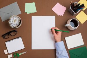 A hand holding a green pencil above a blank paper, surrounded by stationery, a coffee cup, and a small potted succulent.
