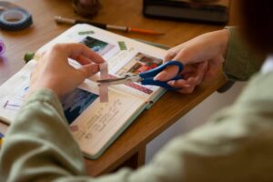 A person with blue scissors cutting decorative tape while working on a scrapbook filled with photos and handwritten notes.