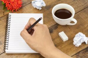 A hand holding a pencil writes in a spiral notebook beside a cup of coffee, an eraser, and crumpled paper on a wooden table.