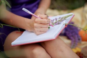 A person writes in a notebook while holding a pink pen, surrounded by flowers and natural elements.