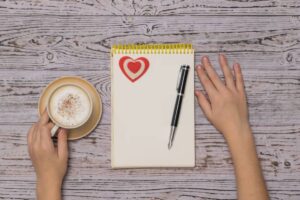 A hand holding a coffee cup next to an open notepad with a heart sticker, alongside a black pen on a wooden table surface.