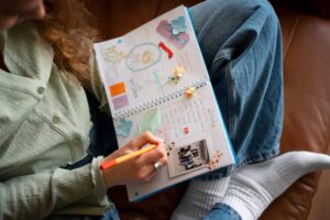 A person writes in a colorful scrapbook decorated with stickers and photos, sitting comfortably with their legs crossed on a couch.