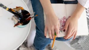 A person's hands hold a sketchbook with pink rose drawings, while a pencil rests in their grip, surrounded by art supplies.