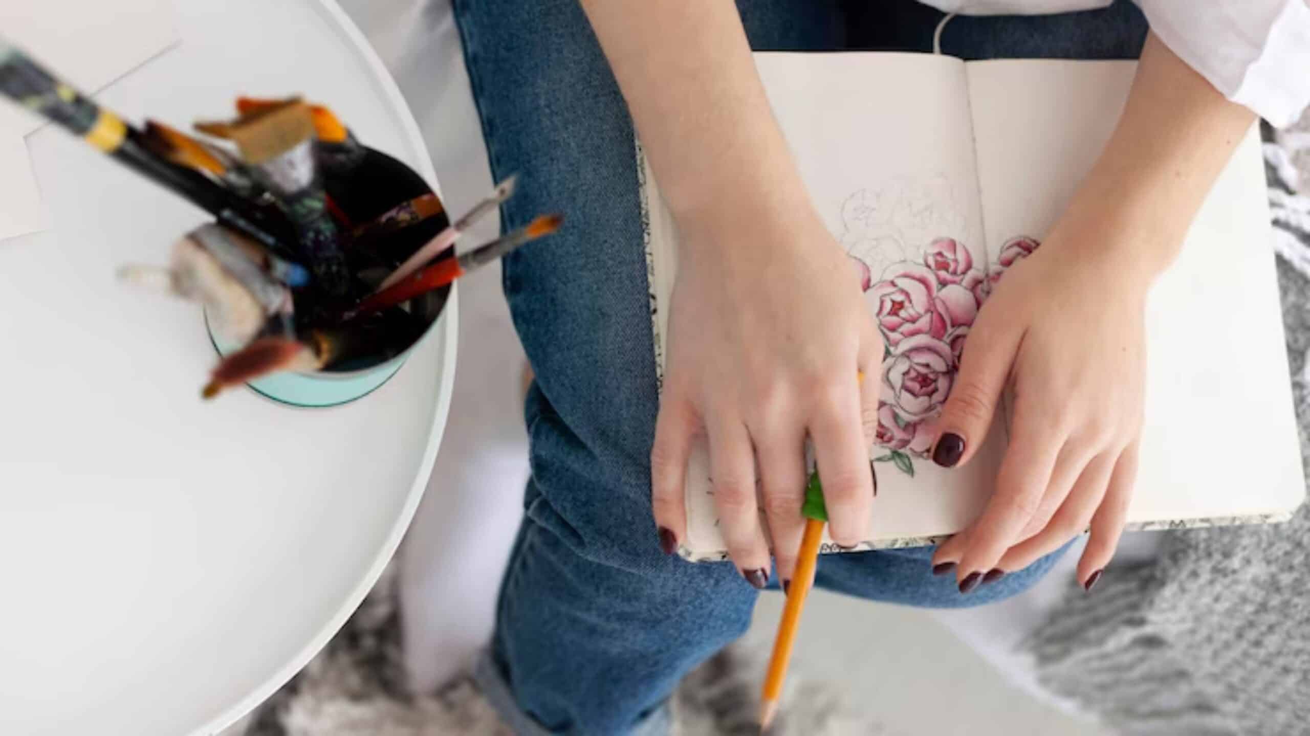 A person's hands hold a sketchbook with pink rose drawings, while a pencil rests in their grip, surrounded by art supplies.