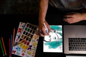 A person drawing on a tablet with an illustration of a woman, surrounded by watercolor paints and colored pencils.