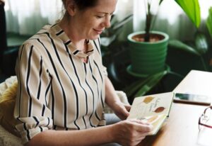 A person wearing a striped blouse sits at a wooden table, examining a notebook with pressed leaves and handwritten notes.