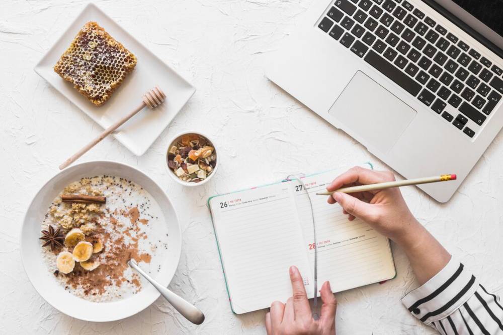 A person writing in a planner while enjoying a breakfast bowl with bananas, honeycomb, and a laptop nearby.