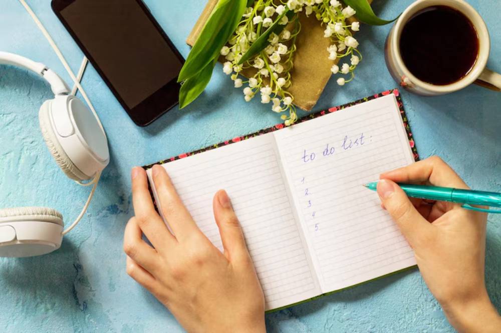 A person's hand writes a to-do list in a notebook, surrounded by headphones, a smartphone, coffee, and flowers on a blue surface.