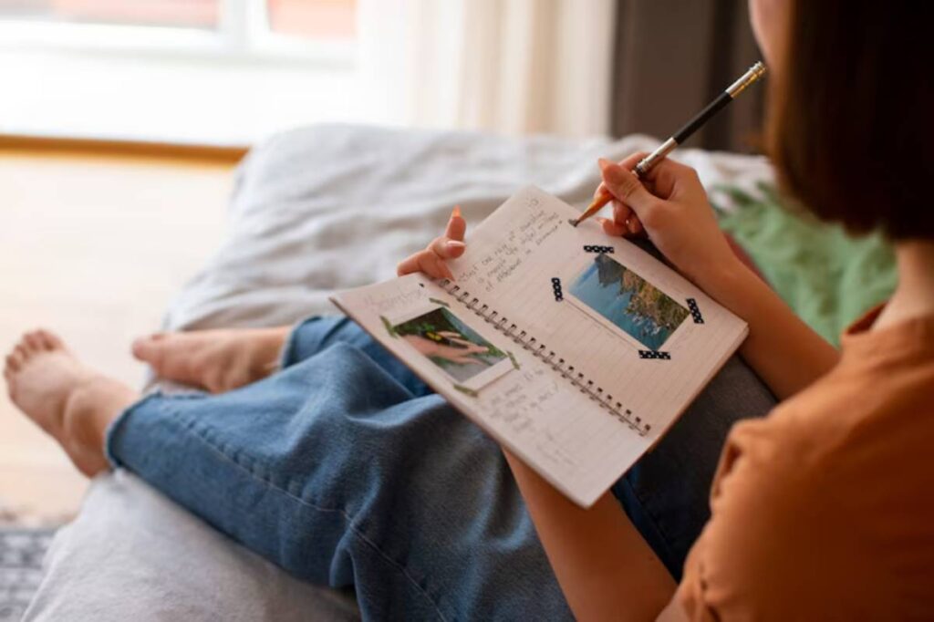 A woman sits on a bed, writing in a notebook adorned with photos and notes, capturing memories in a cozy, sunlit room.