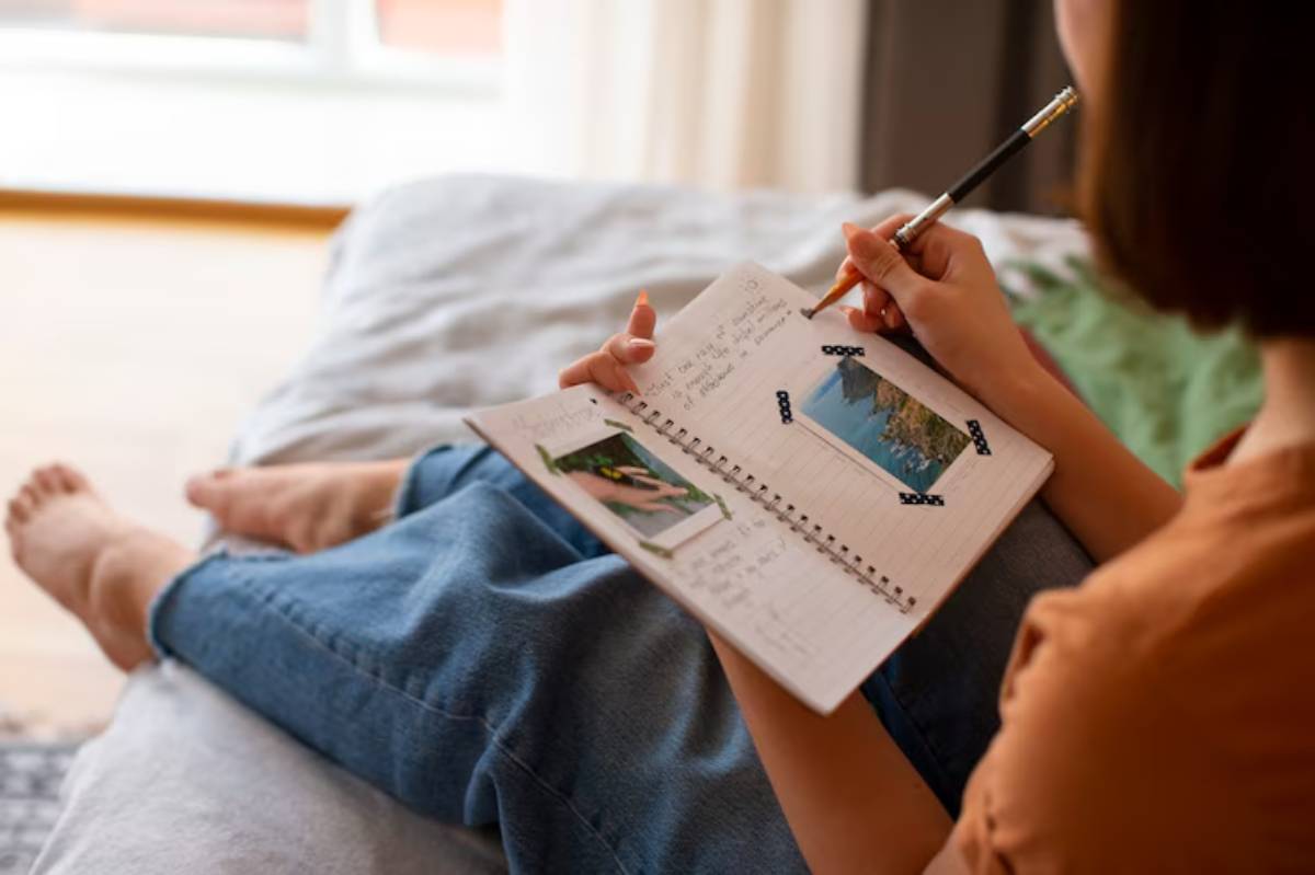 A woman sits on a bed, writing in a notebook adorned with photos and notes, capturing memories in a cozy, sunlit room.